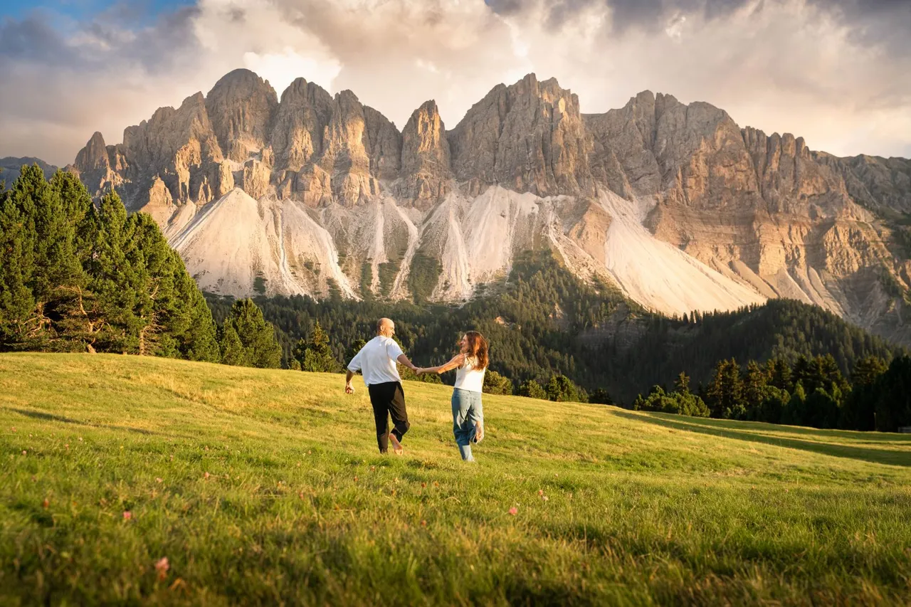 Sarah und Lorenz laufen Hand in Hand über eine Almwiese vor den Dolomiten
